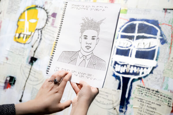 Gros plan sur deux main qui tiennent un cahier avec un dessin en relief d'un portrait de buste de Basquiat. Au fond, une partie d'une oeuvre de peinture et collage de l'artiste.